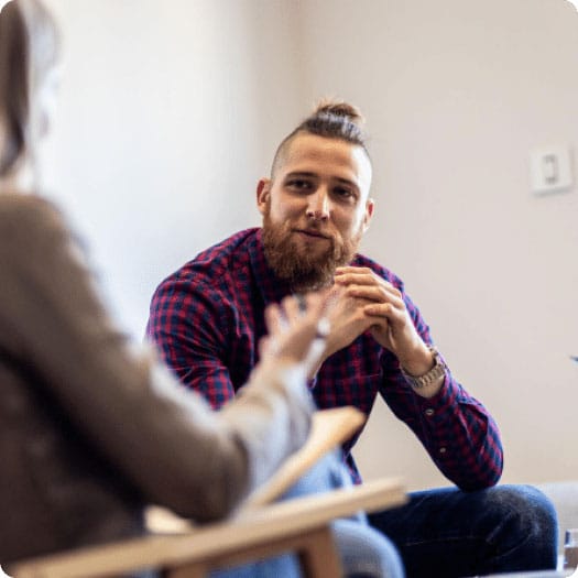 Man with a beard and tied-up hair wearing a red plaid shirt, attentively listening to a woman during a conversation in a room.