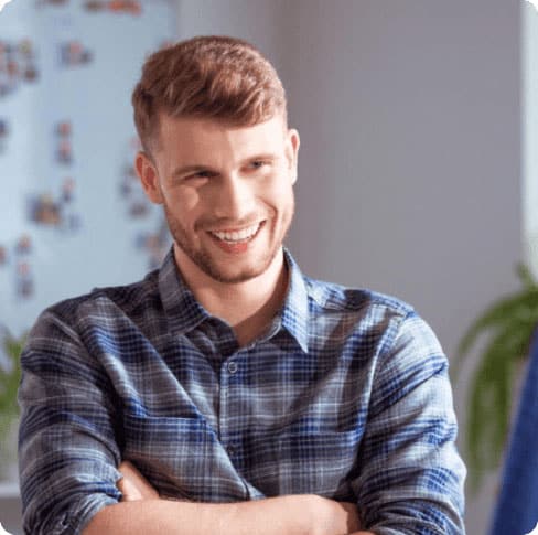 Smiling young man with short brown hair wearing a blue plaid shirt, standing with arms crossed in a well-lit room.