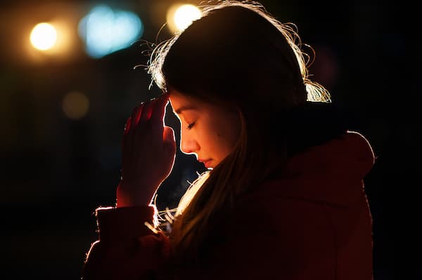 A side profile of a woman with her eyes closed, holding a hand up to her forehead in a pensive or distressed gesture. She is illuminated by a warm rim light against a dark background with soft, blurry streetlights.