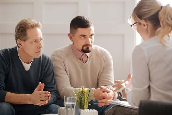 Two men sit on a couch, listening closely to a female therapist who is facing them with her back partially to the camera. The man on the left wears a dark grey sweater with his hands clasped, while the man on the right wears a beige knit sweater over a pink button-down shirt and has a goatee.
