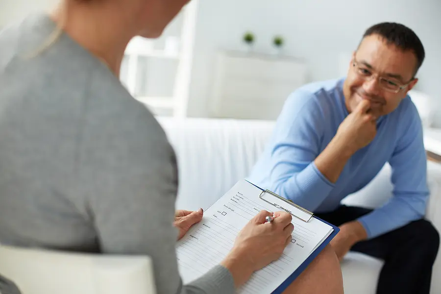 An over-the-shoulder view of a clinician holding a clipboard and writing notes with a pen during an assessment. In the blurred background, a male client wearing a light blue long-sleeve shirt and glasses looks toward her with a slight smile, resting his hand near his chin.