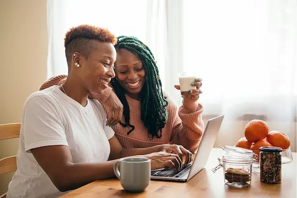 Two Black women sit together at a wooden table, smiling as they look at a laptop screen. One woman with short, reddish-brown hair types on the keyboard, while the other woman with long green braids sits close behind her, holding a small glass of milk. A mug, jars of cookies, and a bowl of oranges rest on the table nearby.
