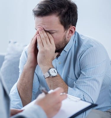 A man wearing a light blue button-down shirt and a silver wristwatch sits with his head down, covering his face with both hands in an expression of distress or exhaustion. In the blurred foreground, a therapist holds a clipboard and a pen, taking notes during the session.
