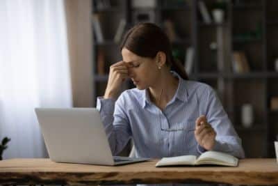 A woman in a striped button-down shirt sits at a wooden desk with a laptop and an open notebook, rubbing the bridge of her nose in frustration or fatigue. She holds a pair of glasses in her other hand, with a blurred bookshelf visible in the background.