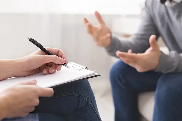 A close-up view focusing on a therapist’s hands holding a black pen over a blank clipboard. In the blurred background, a client wearing a grey sweater gestures with their hands while speaking during a therapy session.