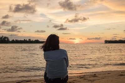A person with shoulder-length dark hair is seen from behind, sitting on a sandy beach and looking out at a scenic sunset over the water. The sky is filled with soft orange and grey clouds, and dark trees line the distant shoreline.