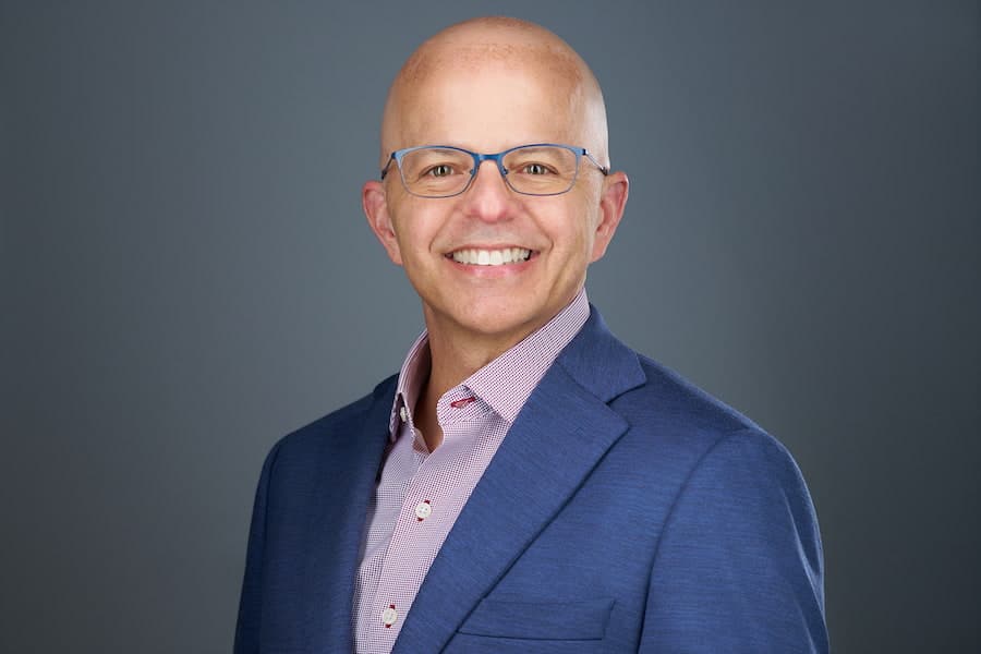 A studio headshot of Dr. Junco smiling against a solid dark gray background. He is wearing blue-framed glasses, a blue blazer, and a pink and white patterned button-down shirt.