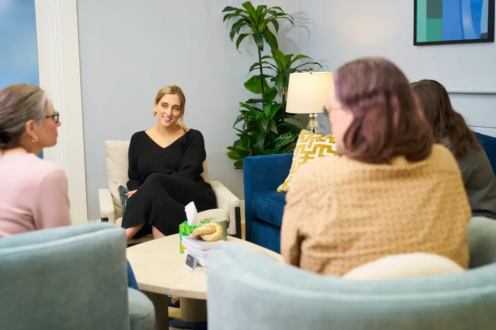 Whitney Polich, wearing a black long-sleeved shirt, sits in an beige armchair and smiles while listening during a group session. Three other people are seated in front of her, with two seen from behind in the foreground, in a cozy office setting with a blue couch and a tall green plant.