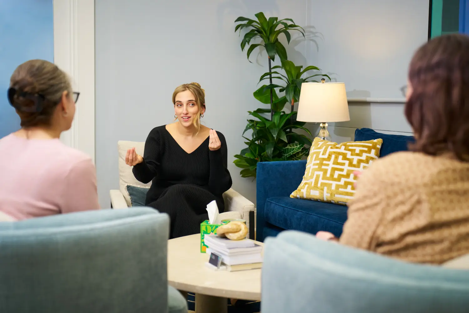 Whitney Polich, a woman with her hair tied up in a bun and wearing a black long-sleeved shirt, sits in an armchair and gestures with both hands while speaking. Two people are seated in front of her with their backs to the camera, listening to her in a cozy office setting that features a blue couch, a patterned yellow pillow, and a large green plant.