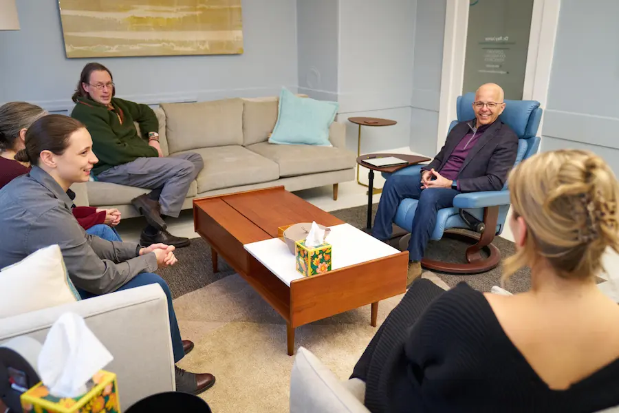 Dr. Junco, wearing glasses, a dark blazer, and a purple shirt, sits smiling in a blue armchair during a group therapy or discussion session. Several participants are seated around a wooden coffee table on couches and chairs, listening and engaging in the conversation within a modern office space.