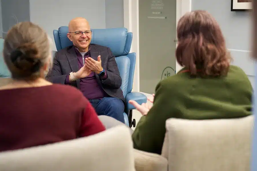 Dr. Junco, a man wearing glasses, a dark blazer, and a purple shirt, sits smiling in a blue armchair while clapping his hands together during a session. Two people are seated in front of him with their backs to the camera, listening to him in a professional office setting.