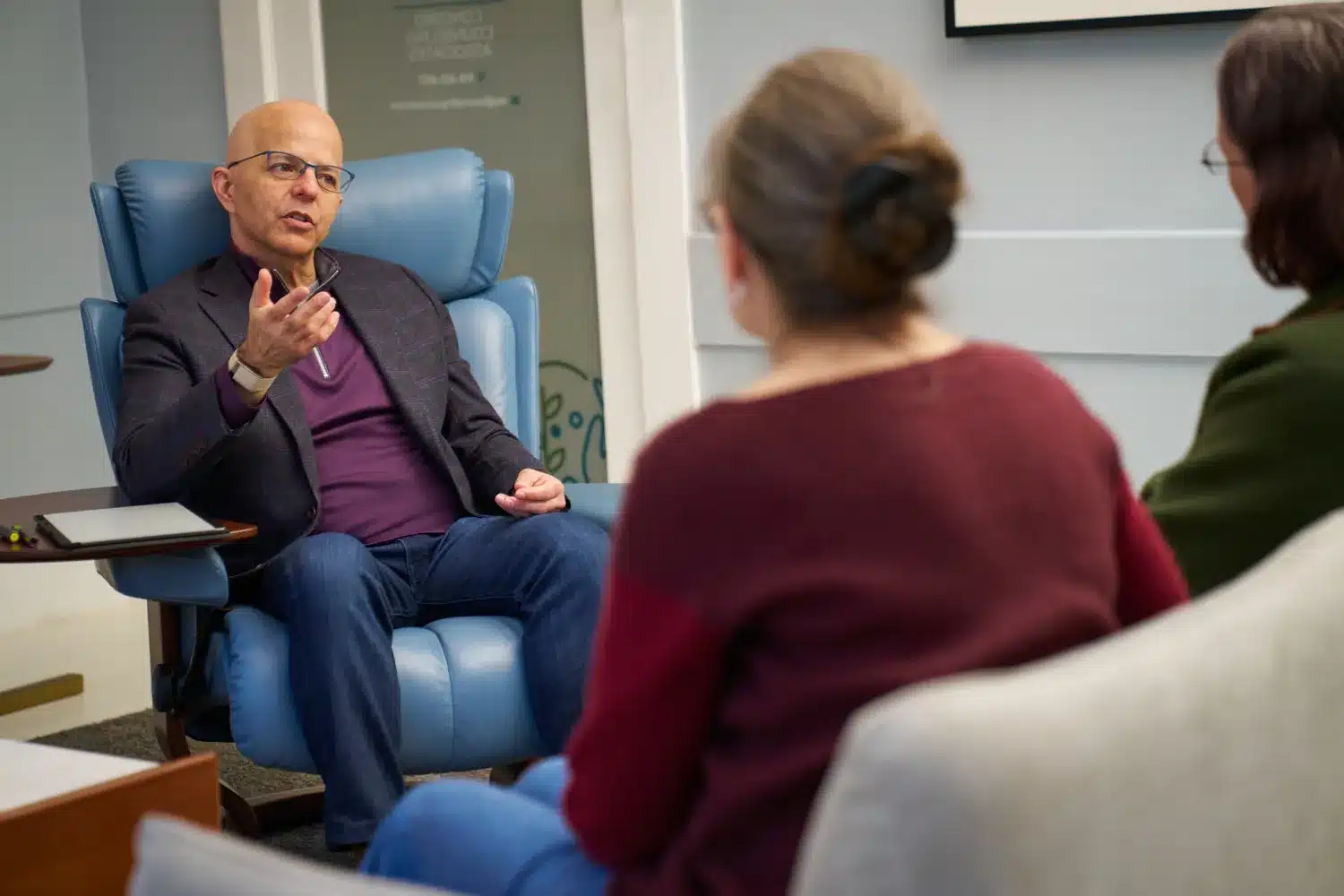 Dr. Junco, a man wearing glasses, a dark blazer, a purple shirt, and jeans, sits in a blue armchair and gestures with his hand while speaking during a session. Two people, seen from behind with their backs to the camera, sit on a couch in the foreground listening to him in a professional office setting.