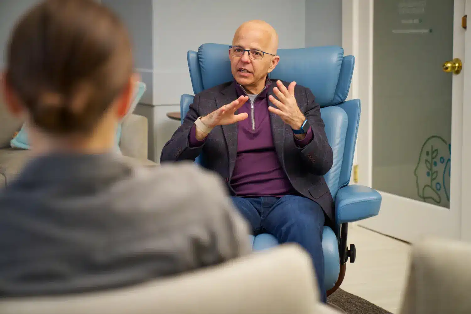 Dr. Junco, a man wearing glasses, a dark blazer, and a purple shirt, sits in a blue armchair and talks with his hands raised. A person is seen from behind in the foreground listening to him.
